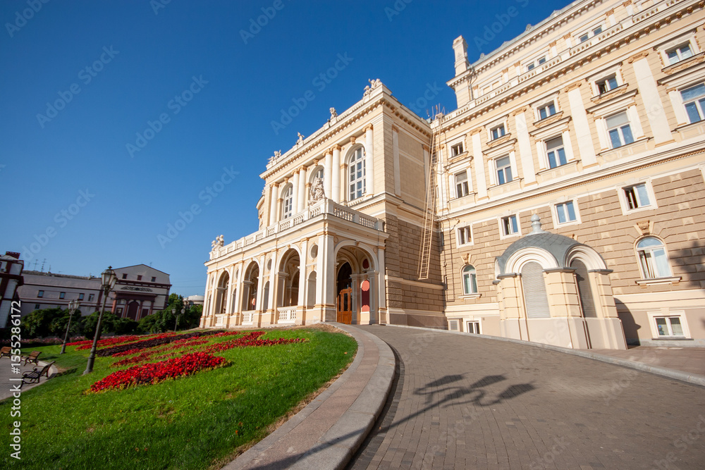 Fototapeta premium Grand Opera and Ballet Theatre building with classical architecture, surrounded by vibrant red flowers and a lush green lawn under a clear blue sky, showcasing culture and heritage