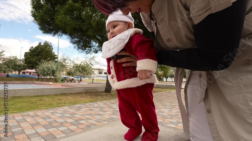 Happy baby boy wearing a Santa Claus costume practicing his first steps, supported by his aunt in a park