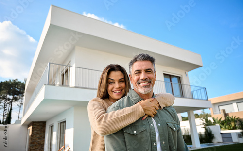 Happy older couple new home owners standing outside constructed or sold house. Smiling villa property buyers investors, affectionate middle aged man and woman in love hugging outdoors while moving in.