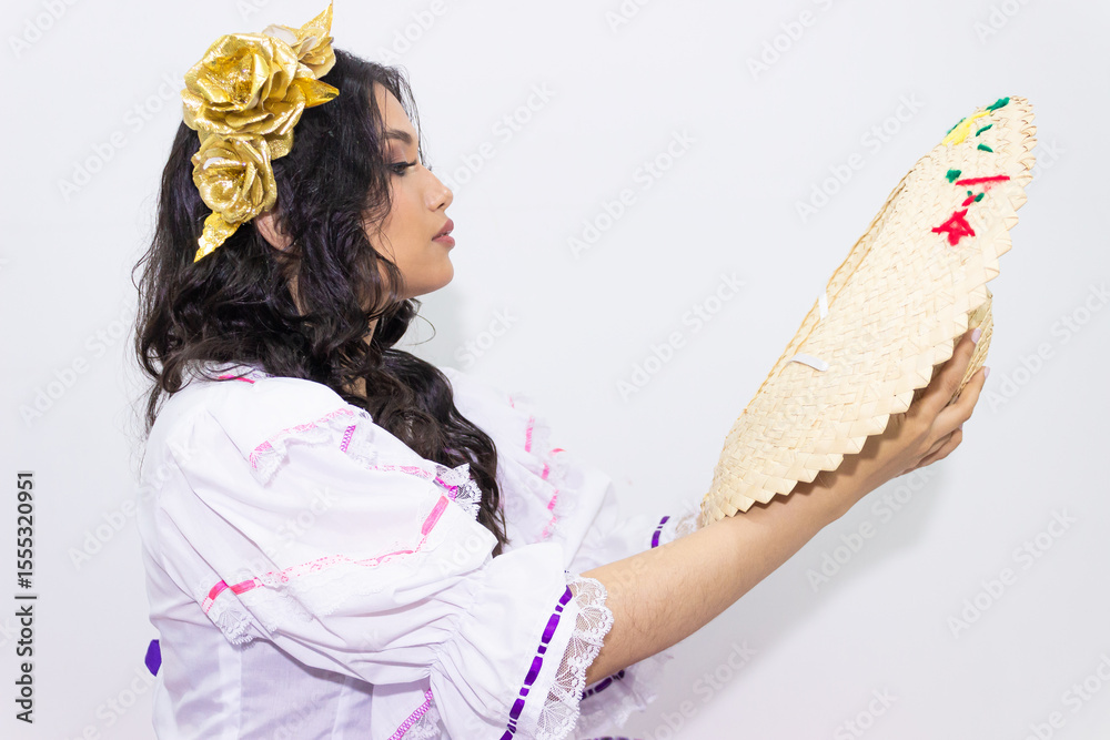 Naklejka premium A young Hispanic woman wearing a traditional costume is looking at a sombrero while holding it in her hands, against a white background. Colombian culture concept