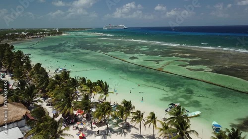 Aerial drone view of tropical beach with palm trees, crystal-clear turquoise waters, and a cruise ship in the distance. Paradise coastal scene ideal for travel and vacation concepts.