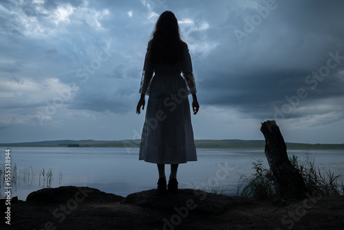 Mysterious woman in white dress standing by a dark lake under stormy sky