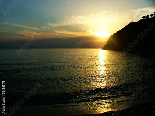 Breathtaking sunset over the Calabrian sea, with vibrant hues of orange, gold illuminating the sky. The calm water reflects the warm colors, while the rugged cliffs of Calabria.