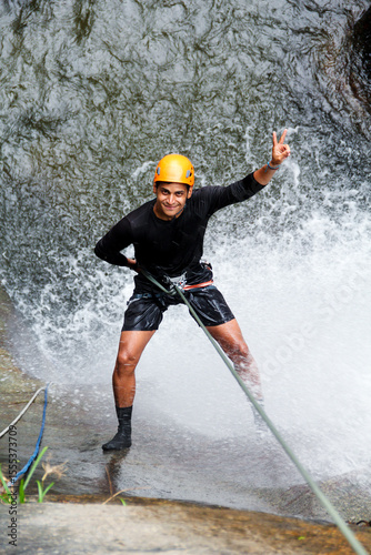 Latin american young man canyoneering and rappelling down a stunning waterfall, flashing a victory sign with his hand, embodying adventure and excitement