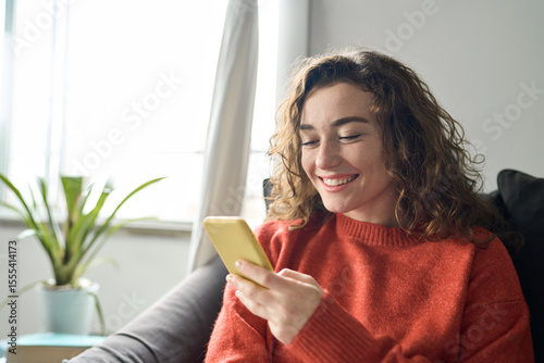 Happy girl holding smartphone, looking at cellphone doing online shopping in mobile apps. Smiling young woman sitting on couch using cell phone ordering ecommerce products or checking social media.