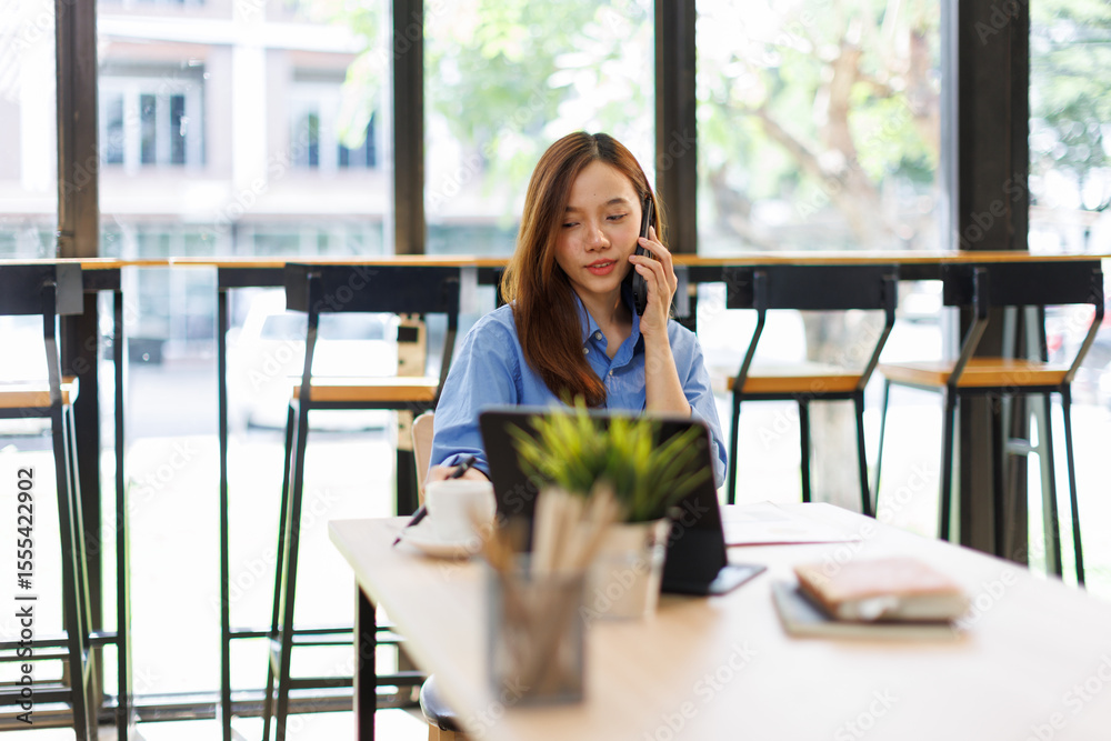 Fototapeta premium Smiling asian woman writing notes while making phone call and using laptop at workplace. 