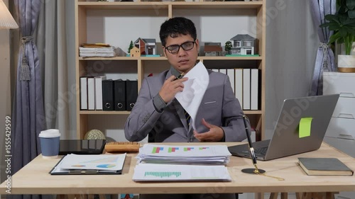 Asian businessman searches documents at night in office with computer showing stock market charts to analyze company finances and taxes, working overtime after work