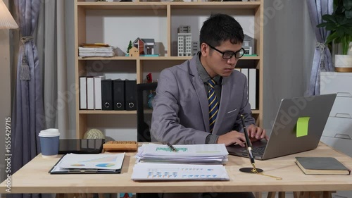 Asian businessman searches documents at night in office with computer showing stock market charts to analyze company finances and taxes, working overtime after work