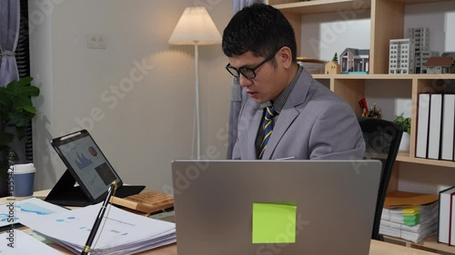 Asian businessman searches documents at night in office with computer showing stock market charts to analyze company finances and taxes, working overtime after work