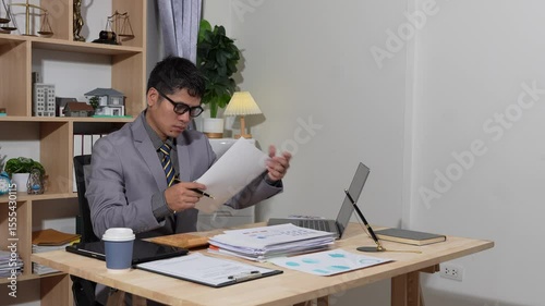 Asian businessman searches documents at night in office with computer showing stock market charts to analyze company finances and taxes, working overtime after work