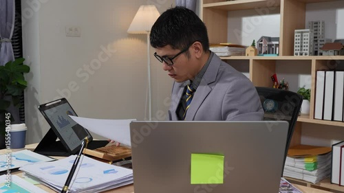 Asian businessman searches documents at night in office with computer showing stock market charts to analyze company finances and taxes, working overtime after work
