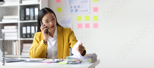 A serious female employee is busy with paperwork and talking on the phone with the boss.
