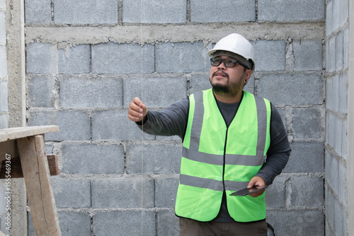 Young Asian engineering worker with safety helmet overweight employee foreman inspect property project on construction site, contractor architect man wearing eyesight glasses holding tablet technology
