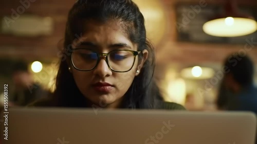 Young woman focused on her laptop in a cozy caf?, with blurred patrons and warm lighting in background