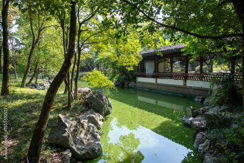 Traditional Chinese pavilion beside a green pond, surrounded by rocks and trees in a classical landscaped park. Classic community public green space or parkland in China.