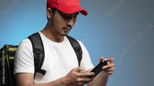 Young man in a red cap using a smartphone, focused on the screen, with a backpack in a studio setting