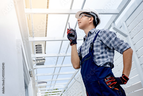 Construction engineer man using walkie talkie to cooperate with the worker team while looking at roof structure. Asian male foreman talking on radio communication equipment at house building site.