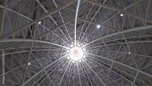 Symmetrical view of a modern architectural ceiling featuring a geometric steel framework and a central skylight. Captured from directly below, showcasing the intricate pattern and industrial design