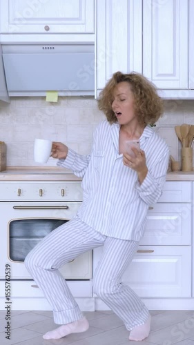 Vertical screen happy woman in pajamas enjoying her morning routine, dancing and singing with a cup of coffee and smartphone in her modern white kitchen