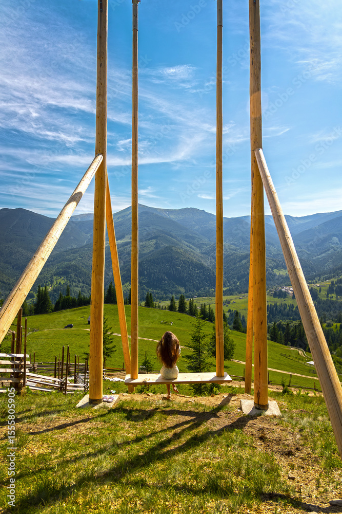 Fototapeta premium Girl on giant swing overlooking mountain valley. Young girl sits on a massive wooden swing, facing a stunning mountain landscape under a bright blue sky in summer.