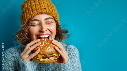 Happy young woman in a mustard beanie and blue sweater enjoying a delicious burger, combining cozy fashion with vibrant summer energy against a blue background