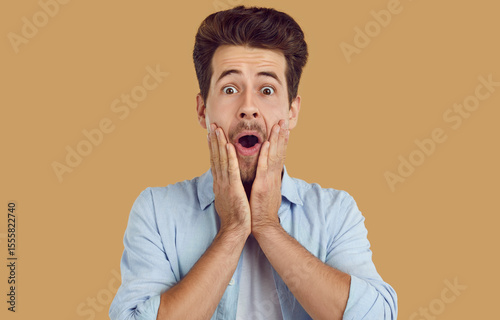 Portrait of young amazed scared man in blue shirt on beige background looking at camera. He touching his surprised face two hands on chin. Wide-open eyes and mouth in horror. Human sincere emotion.