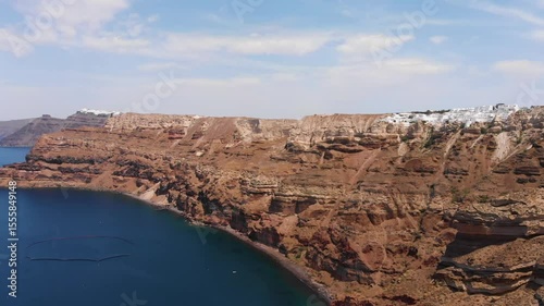 Aerial footage capturing the dramatic volcanic caldera cliffs of Santorini, Greece, with white-washed villages perched atop, overlooking the deep blue waters of the Aegean Sea under a clear sky.