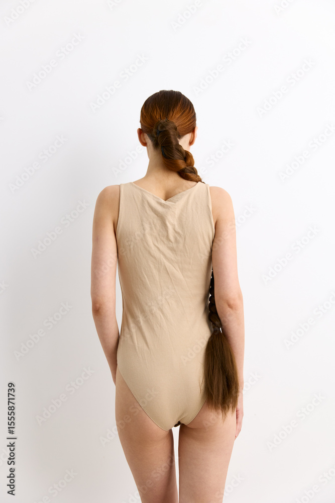 Obraz premium Woman with long braided hair wearing beige bodysuit stands against a white seamless background showing back view with minimalist style and natural lighting.