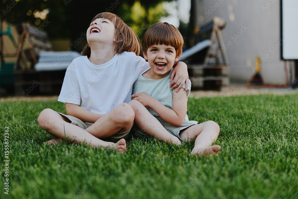 Fototapeta premium Two happy boys laughing together while sitting on the grass, enjoying a sunny day. Their playful expressions capture the innocence of childhood in a natural setting.