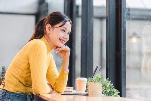 Free Download Young woman in yellow sweater smiling while looking at tablet near window with coffee cup and plant, enjoying peaceful moment indoors - GFXLoad