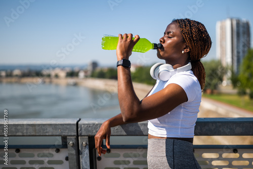 Portrait of young fit African woman in sport clothing with headphones drinking energy drink after sport training on urban city bridge.	