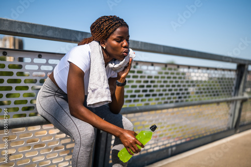 Portrait of exhausted African woman in sports clothing wiping sweat with towel, drinking energy drink and resting after sport training on fence of the city bridge.