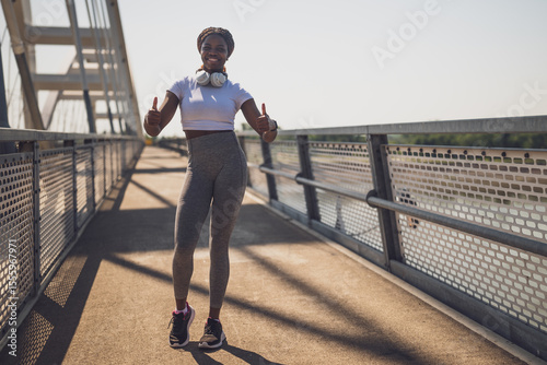 Portrait of happy sporty African woman in sport clothing wearing headphones and showing thumbs up during exercise training on urban city bridge.