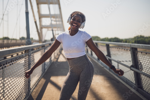 Cheerful African sporty woman in sports clothing enjoying listening to music through headphones during exercise training on urban city bridge.