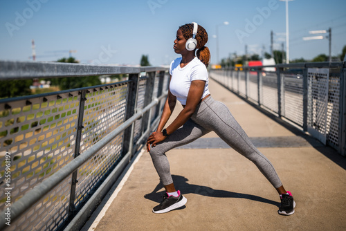 Sporty young African woman in sportswear with headphones enjoying exercising and stretching while standing at the city bridge. She is in the squatting position.	