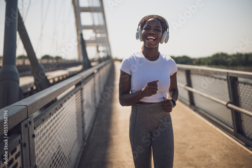 Happy and fit sporty African woman in sportswear jogging and listening music through headphones on urban city bridge.	