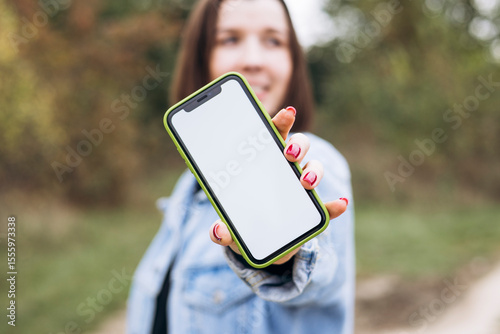 Smiling young woman showing blank smartphone screen to camera outdoors.