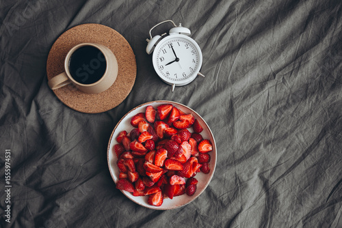 A plate with fresh strawberries, an alarm clock and a cup of coffee in bed.