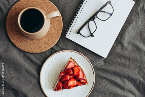 Homemade cheesecake with fresh strawberries and coffee cup on grey bed linen.