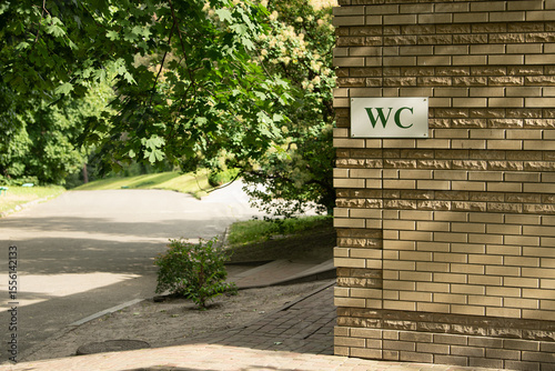A restroom facility marked with a 'WC' sign is situated beside a winding pathway in a park. The area is shaded by trees, creating a serene environment for visitors