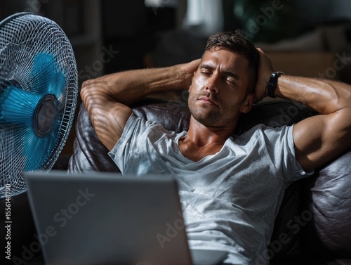 Man in Summer Heat: A man relaxes, cooled by a fan, while working on a laptop, portraying a relatable image of beating the summer heat.