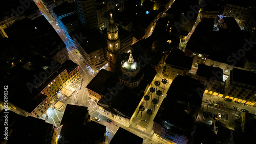 Overhead night aerial of Rapallo Basilica of Saints Gervasius and Protasius with glowing bell-tower and dome framed by palm-lined streets in the old town, Italian Riviera, Liguria, Italy