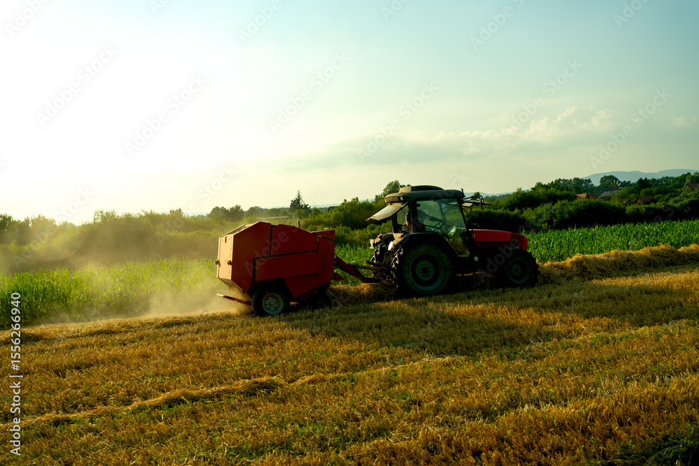Obraz premium Agricultural scene with a red tractor and hay baler operating in a sunset field during harvest season