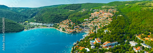 Panoramic aerial view of a coastal town of Rabac with red-roofed buildings nestled between lush green hills and turquoise waters of the Adriatic Sea. Croatia