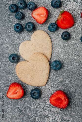 Heart shaped cookies surrounded by fresh strawberries and blueberries on gray surface