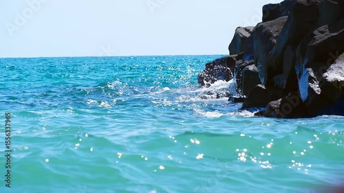 Sea waves crash against the rocky shore. Turquoise water splashes against dark rocks, runs up against rocks. Bright sunlight. Slow motion, soft focus. Nobody.