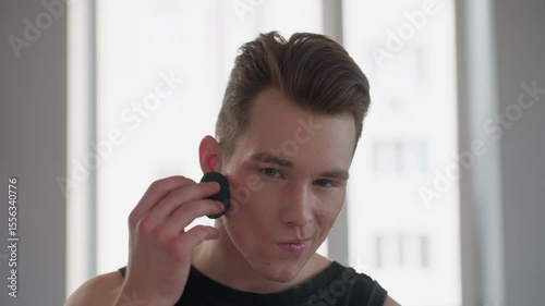 handsome young man in sleeveless signet with neck tattoo sits before bright window mirror, holds black makeup sponge, gracefully dabs powder on cheeks, exudes style and confidence