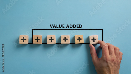 Conceptual image of value added with wooden blocks featuring plus signs arranged in a line and a hand adding another block