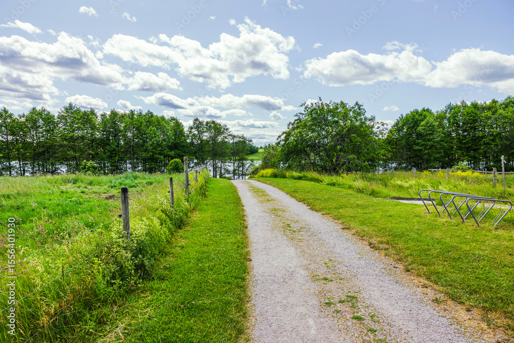Naklejka premium Gravel path leading to lake through green field with bike racks and fence under blue sky with clouds. Sweden.