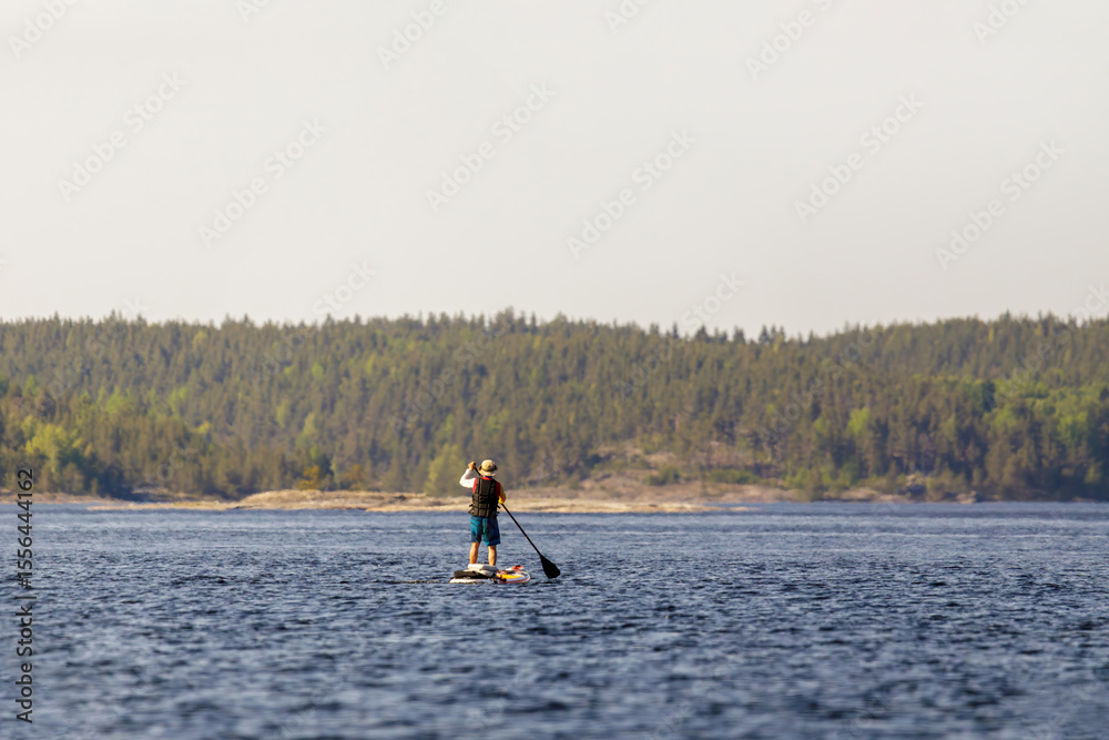 Naklejka premium A person paddleboards across a peaceful lake, pausing to take in the scenic view of lush forest-covered hills.